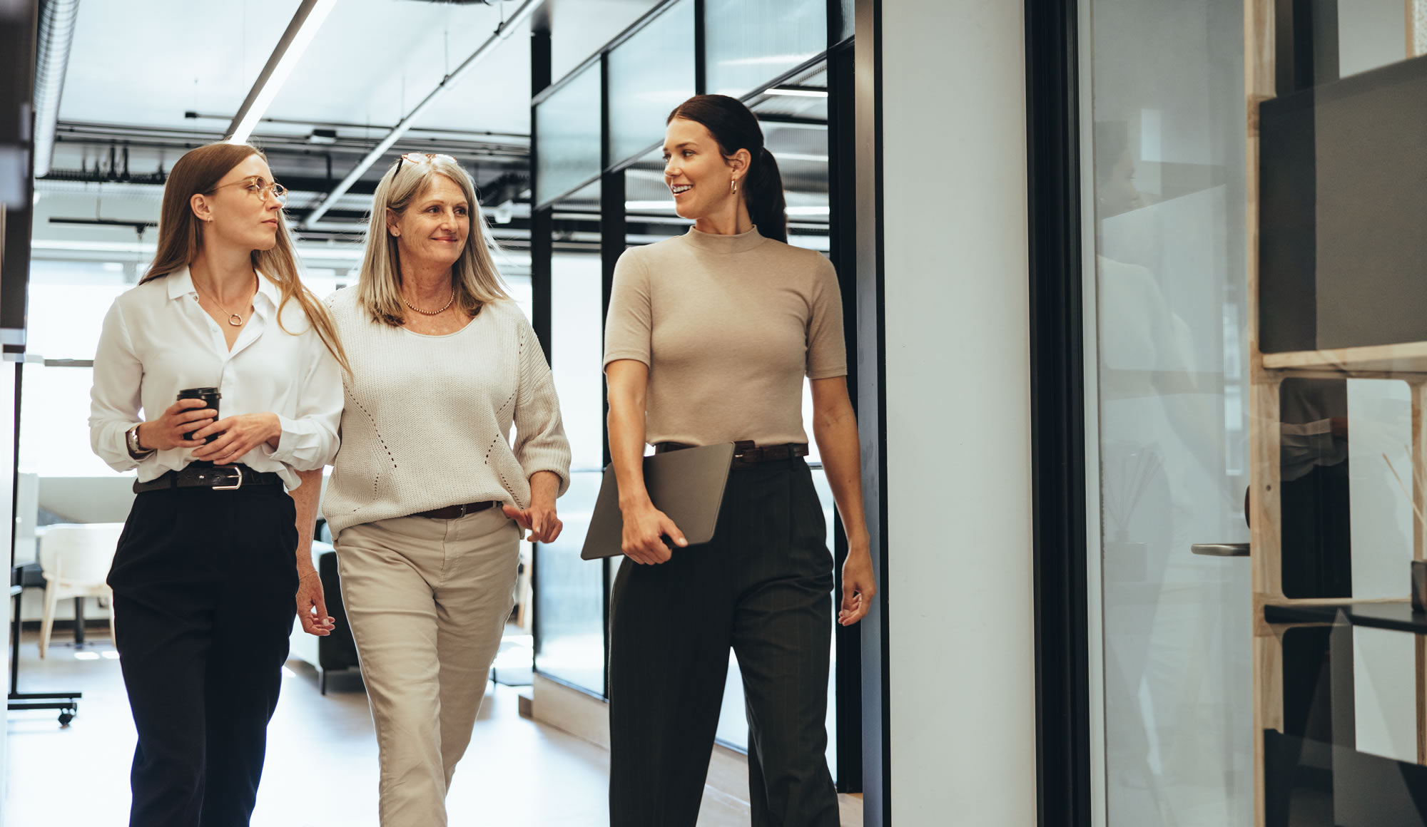 Three women from different generations walking and talking in a modern workplace